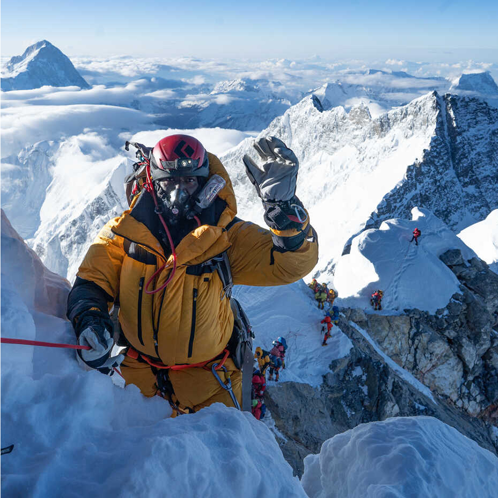 A climber in a yellow down suit, red helmet and oxygen mask, one gloved hand raised, on a snow ridge high above a layer of cloud. Behind and below, a line of other climbers is strung out along the ridge. Snow peaks extend to the horizon.