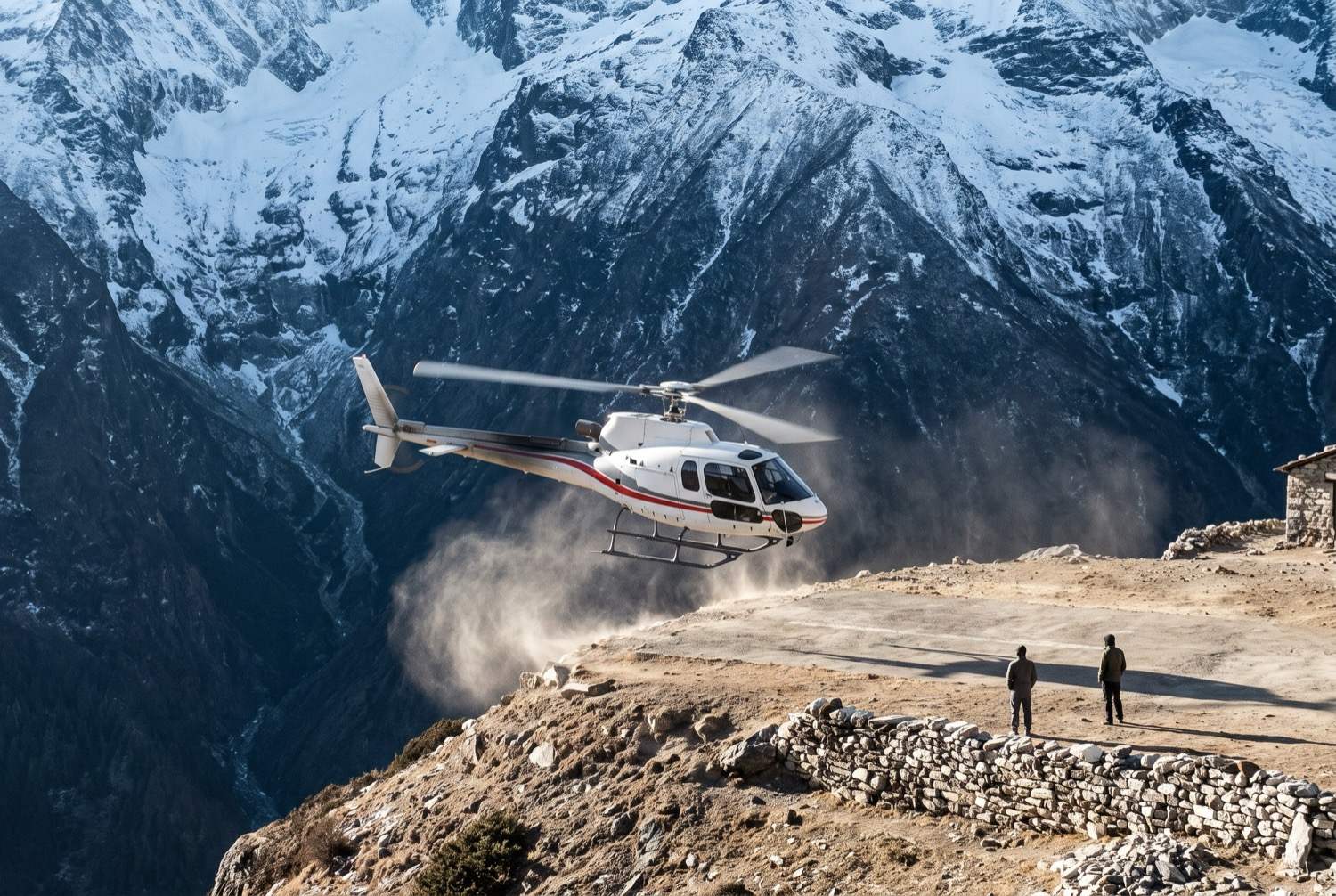 A single-engine helicopter on a short shelf airstrip cut into a mountainside, rotor turning and dust lifting, the ground dropping away into a deep valley beyond the apron edge and an enormous wall of snow and rock rising behind.