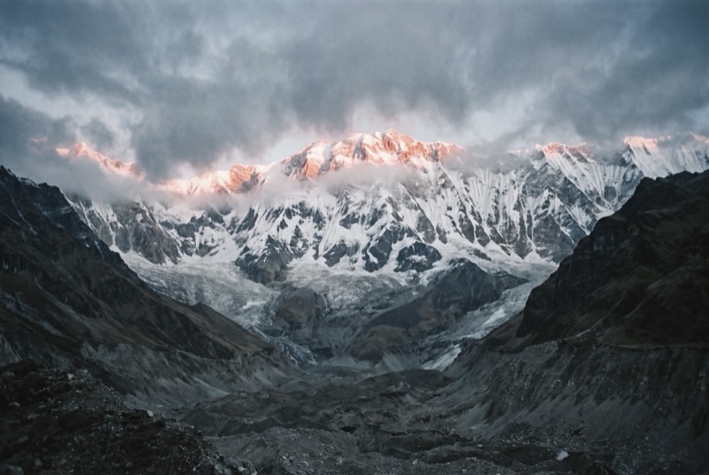 The Annapurna Sanctuary at first light and empty of people: a grey moraine basin ringed by fluted ice walls, with sunrise reaching only the highest snow.