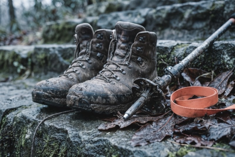 Mud-caked leather trekking boots and the steel tip of a trekking pole on wet stone steps scattered with fallen rhododendron leaves.
