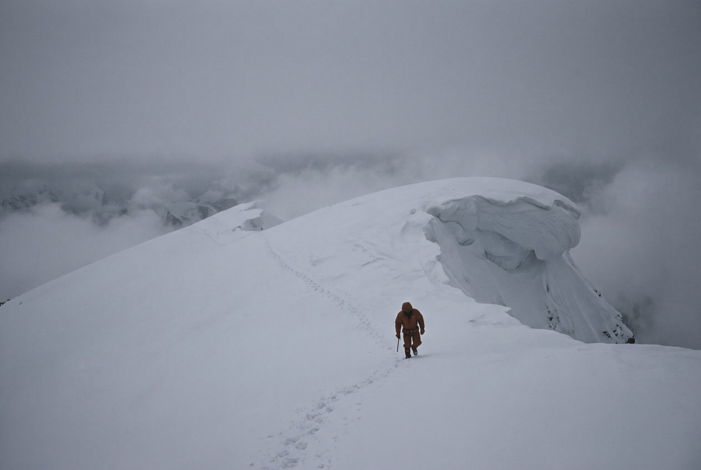A lone climber in an orange down suit far out along the corniced summit ridge of Baruntse, a single line of footprints behind them in flat grey light