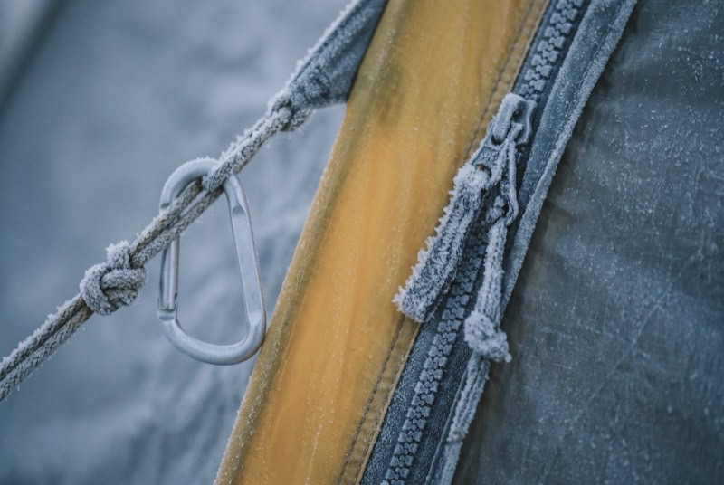 A tent zip frozen shut under hoar frost, a plain carabiner and a knotted guy line clipped alongside it on yellow nylon.