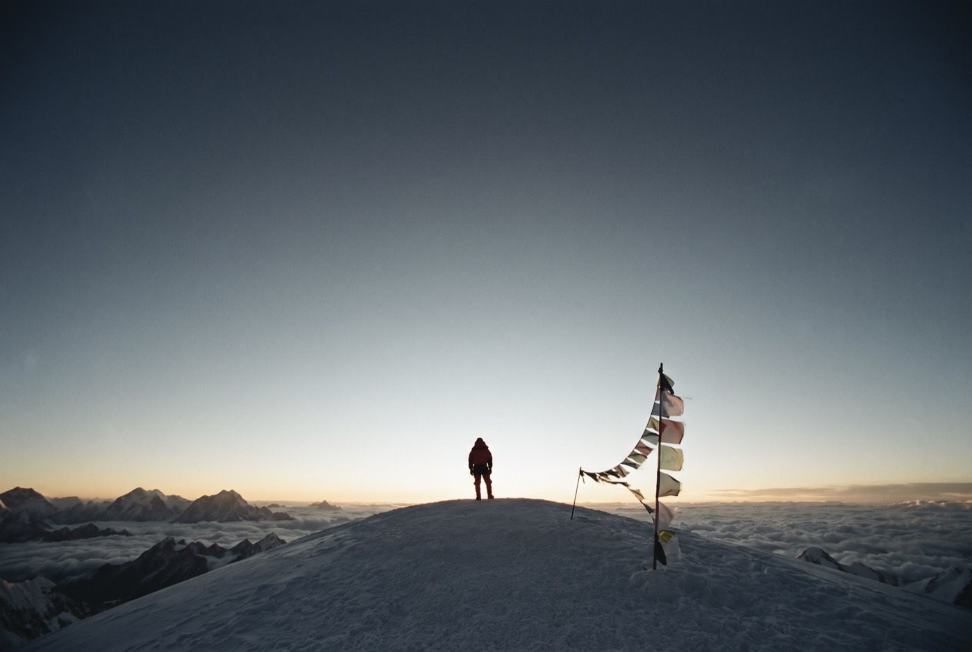A single climber standing on the summit of Everest at sunrise beside a string of prayer flags, a sea of cloud below
