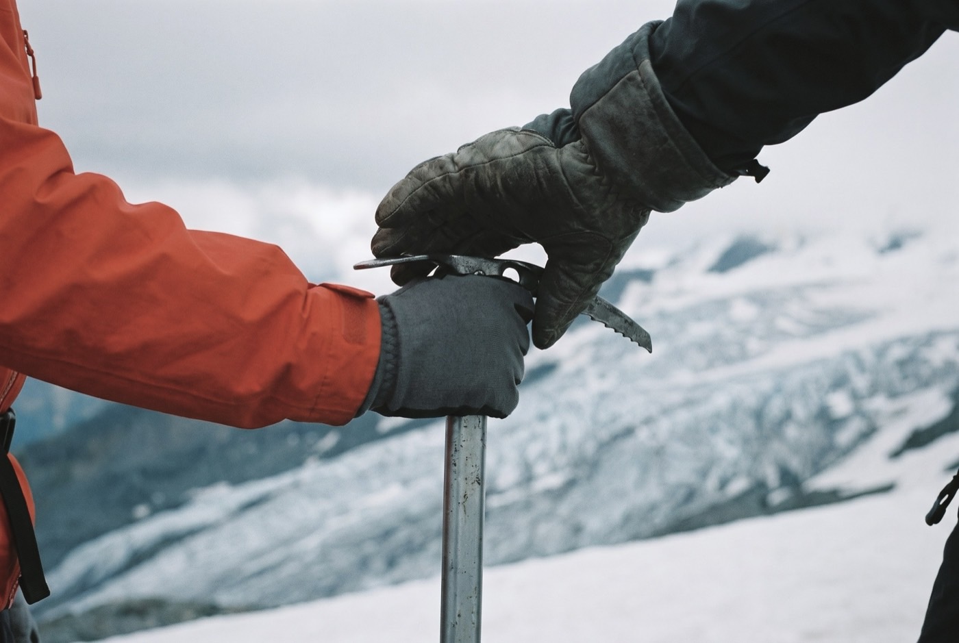 An instructor's gloved hand closes over a client's grip on the head of an ice axe, correcting it, with the glacier falling away out of focus behind.