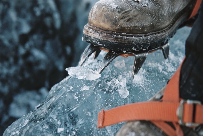 Crampon front points biting into hard blue glacial ice, chips of ice thrown around the steel and an orange strap just in frame.